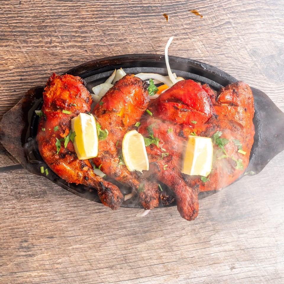 Steaming platter of red grilled chicken pieces garnished with lemon wedges, cilantro, onions, and peppers on a wooden table.