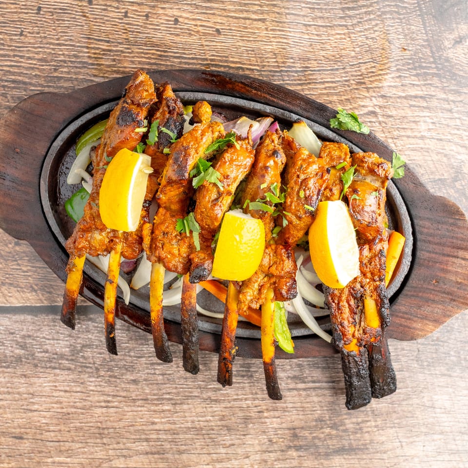 Grilled, spiced meat skewers served on a sizzling platter with lemon wedges, onions, peppers, and fresh cilantro.