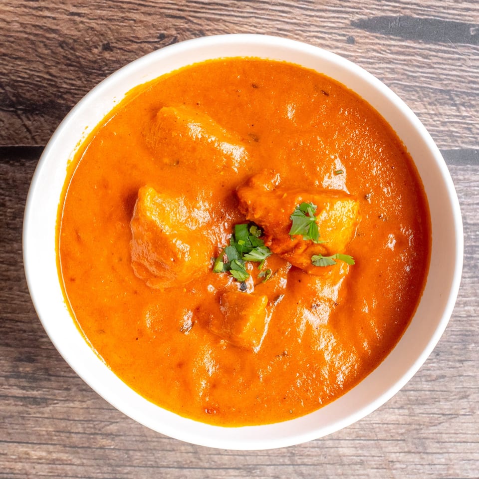 Creamy orange curry with paneer cubes garnished with fresh cilantro, served in a white bowl on a wooden table.