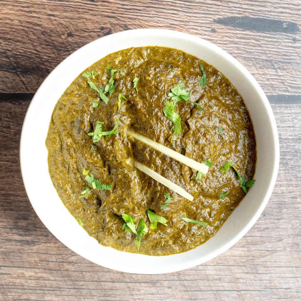 Creamy green curry topped with fresh cilantro and ginger strips in a white bowl on a wooden table.