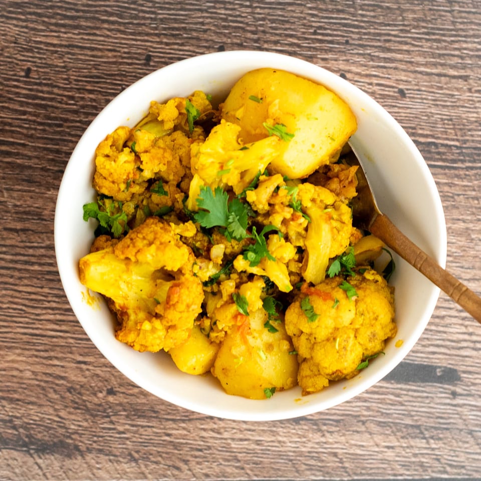 Bowl of golden spiced cauliflower and potatoes garnished with fresh cilantro on a wooden table.